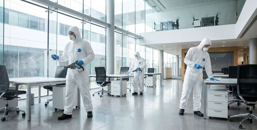 Workers in protective suits disinfecting a modern office with desks and glass walls